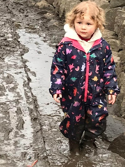 A young girl in a splash suit and wellies walks through a muddy puddle.