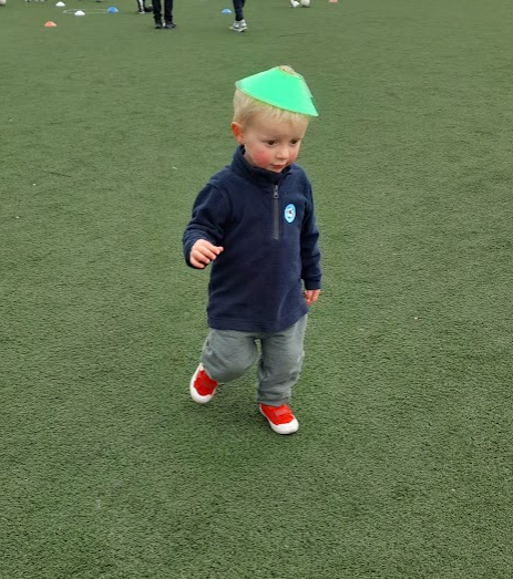 A young boy on an AstroTurf football pitch with a green sports cone on his head.