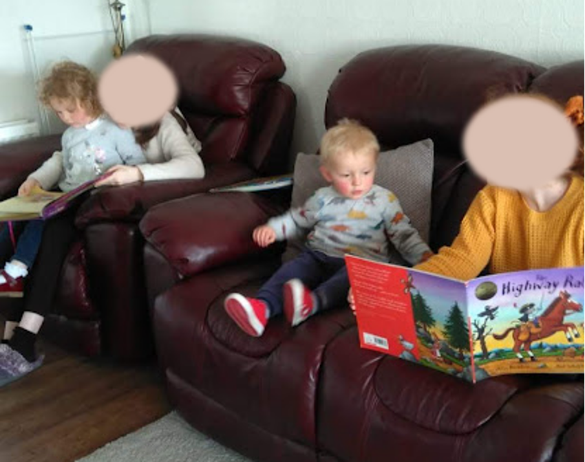 Two teenage girls sat on sofa sharing books with a young girl and toddler boy.