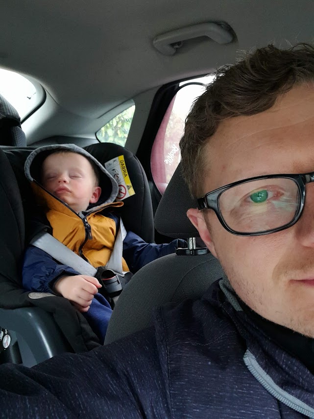 Inside a car, a man wearing glasses looks into the camera and a baby boy sleeps in the back seat.