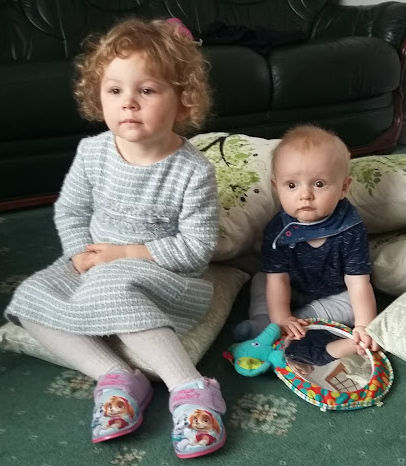 A young girl and baby boy sit on some cushions on a living room floor.