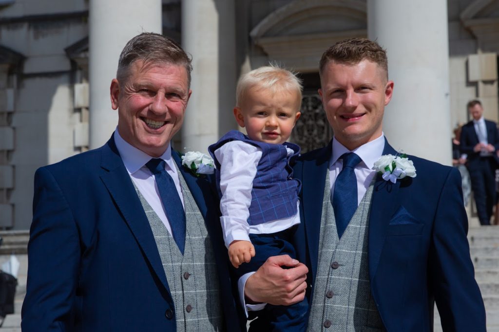 A groom is holding his young son in his arms and is standing next to his father