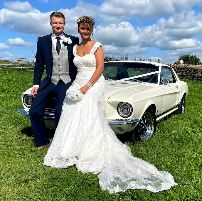 Newly married husband and wife pose while sat on the bonnet of an old Ford Mustang