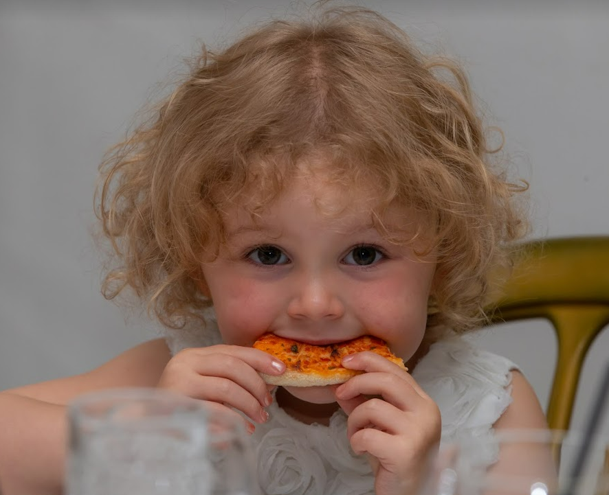 A young girl in a bridesmaid dress eats a slice of pizza.